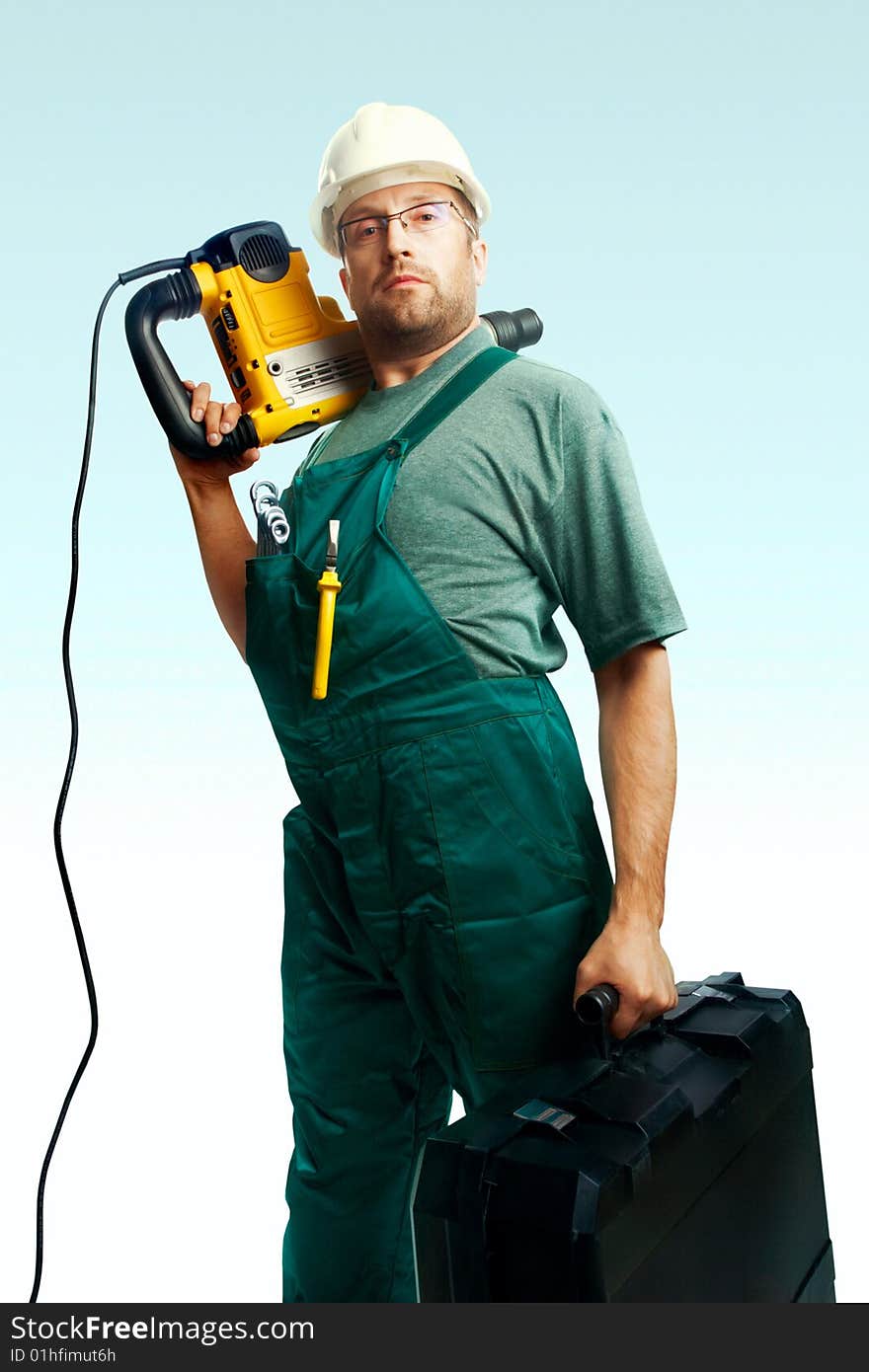 Serious workman in helmet, glasses and overalls hold perforator on the shoulder and big black suitcase in hand over white background. Serious workman in helmet, glasses and overalls hold perforator on the shoulder and big black suitcase in hand over white background