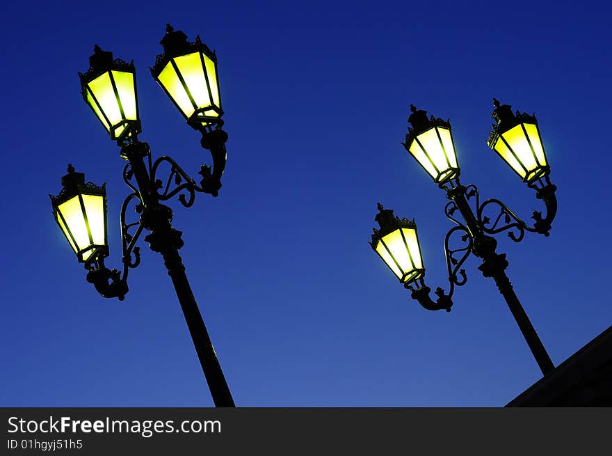 Two sloped, street lights in night dark blue sky. Yellow luminescence. Two sloped, street lights in night dark blue sky. Yellow luminescence.