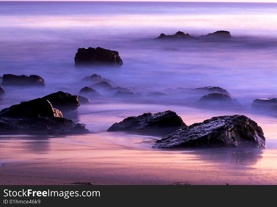 Rocks off the coast of Southern California sit in the mists of the rolling tides at sunset. The beautiful colors of the dusk sky are reflected in the turbulent waters. Rocks off the coast of Southern California sit in the mists of the rolling tides at sunset. The beautiful colors of the dusk sky are reflected in the turbulent waters