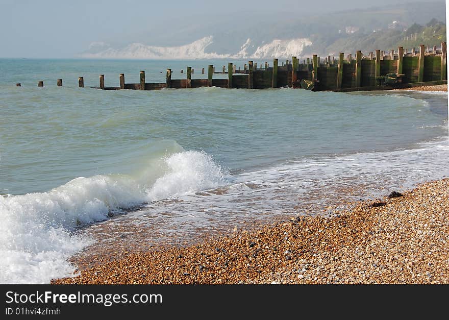 Waves breaking on a pebble beach groynes and cliffs in background.