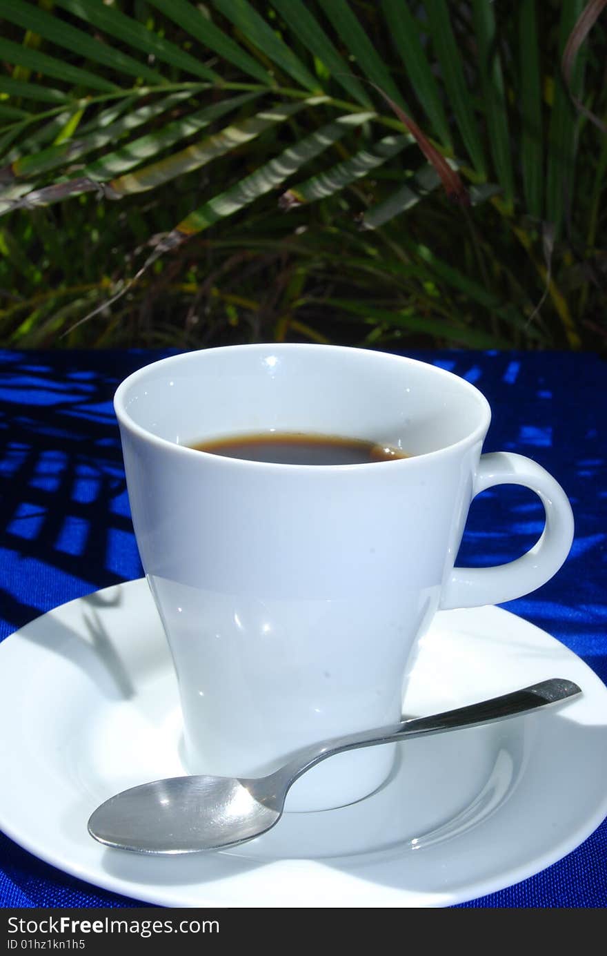 Black coffee in white cup with plate and spoon on blue and palms in the background