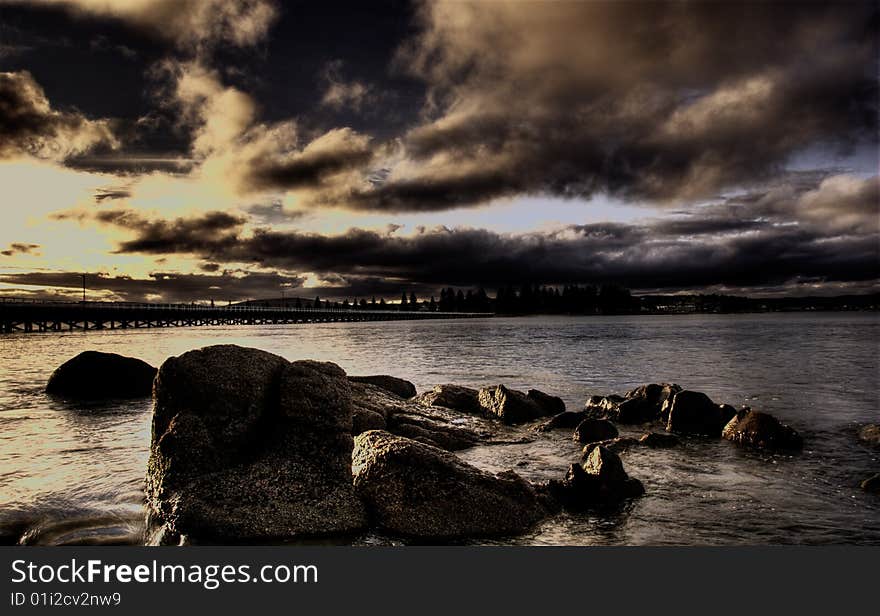 A brooding sunset over the sea with rocks in the foreground and causeway in the background.