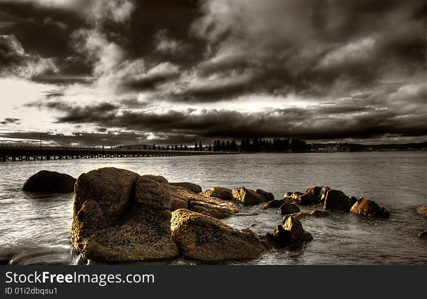 A brooding sunset over the sea with rocks in the foreground and causeway in the background.