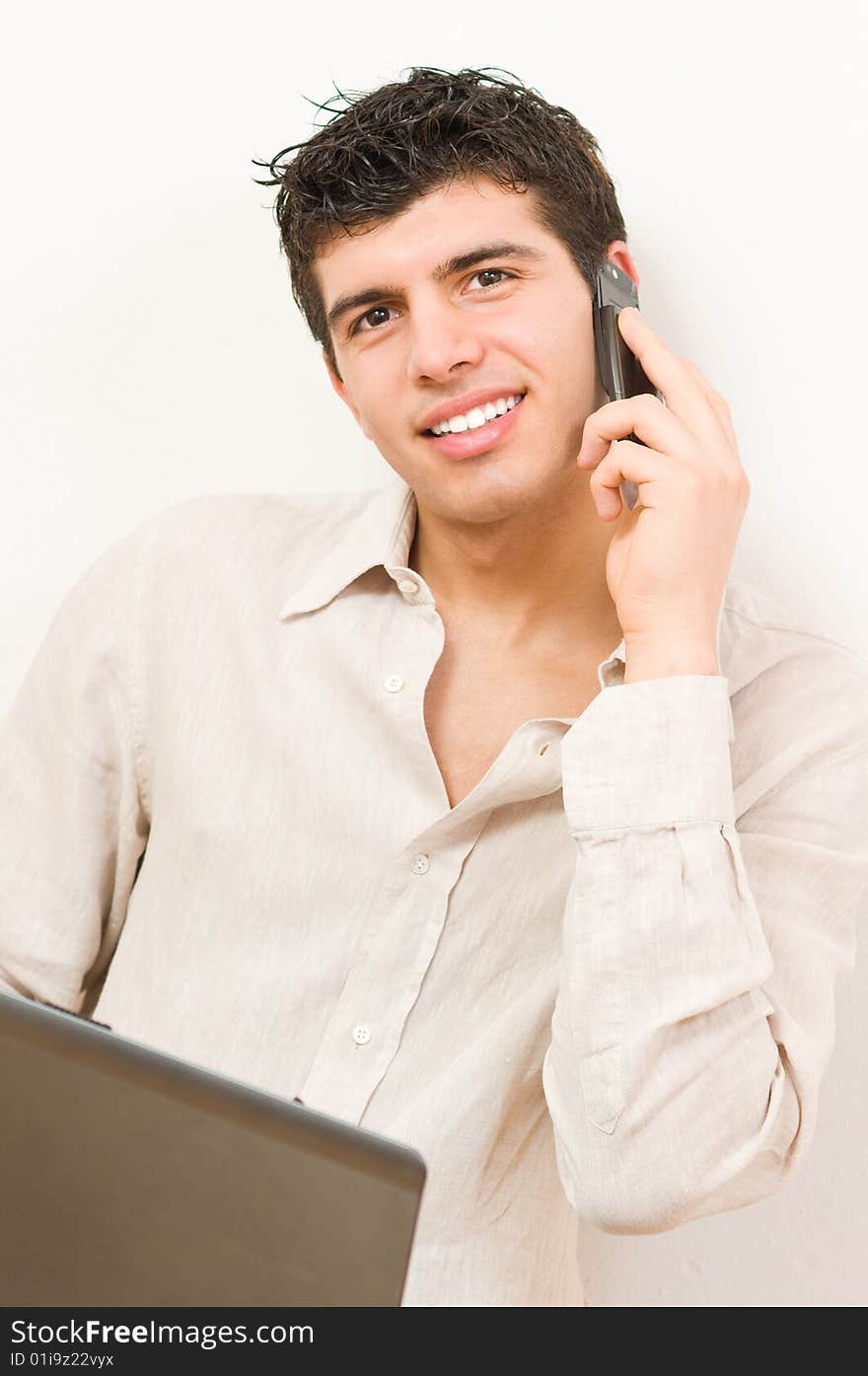 Young man in casual clothing working on his laptop while speaking at mobile phone. Young man in casual clothing working on his laptop while speaking at mobile phone