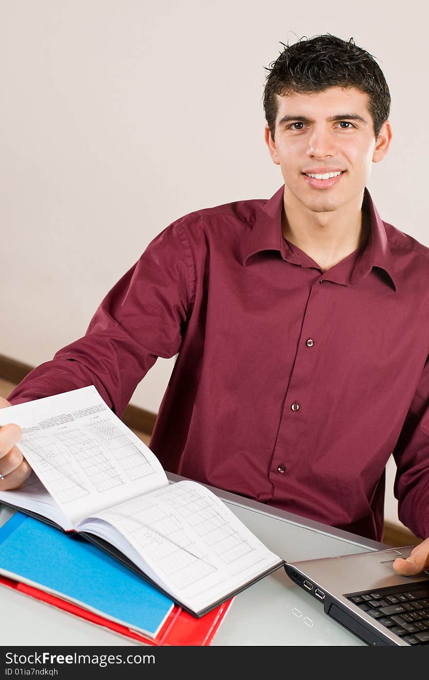 Young man working and smiling at camera with note pad and laptop on desk. Young man working and smiling at camera with note pad and laptop on desk