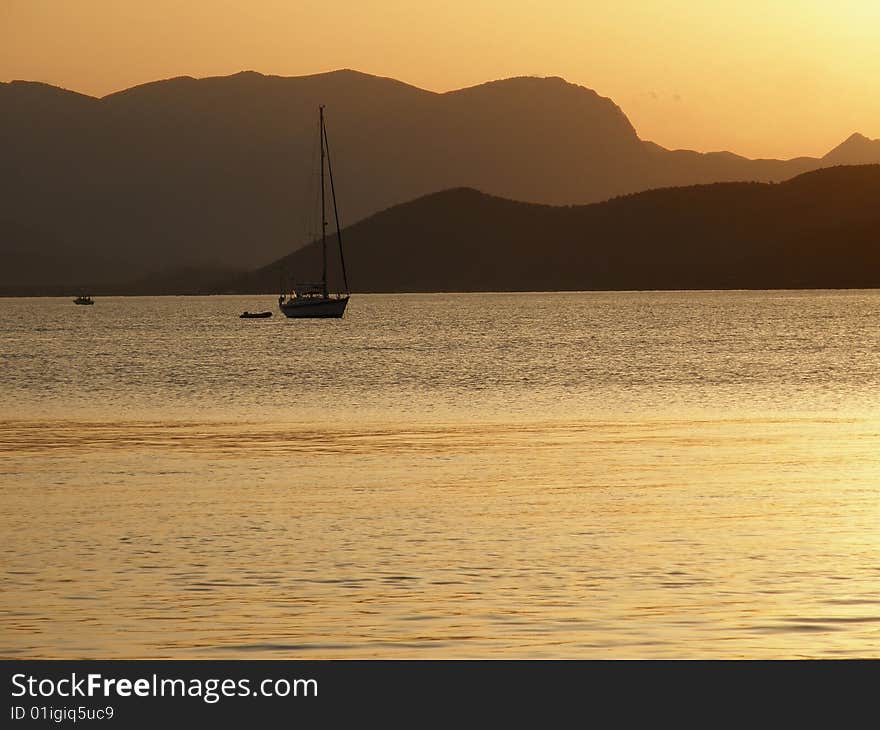 The sun sets over a sailboat on the sea with mountains as a background. The sun sets over a sailboat on the sea with mountains as a background