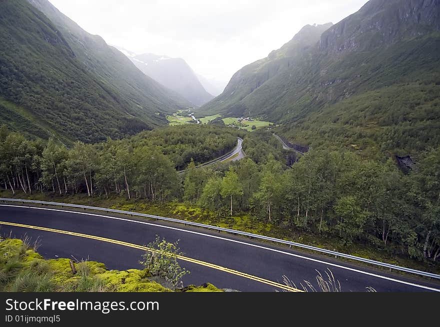 A valley between forest covered mountains. Norwegian landscape on a rainy and misty day. Wet road in the foreground. A valley between forest covered mountains. Norwegian landscape on a rainy and misty day. Wet road in the foreground.