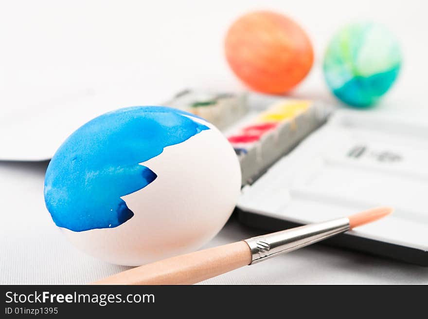 A half painted blue easter egg with brush and watercolor palette and color easter eggs in background. A half painted blue easter egg with brush and watercolor palette and color easter eggs in background.