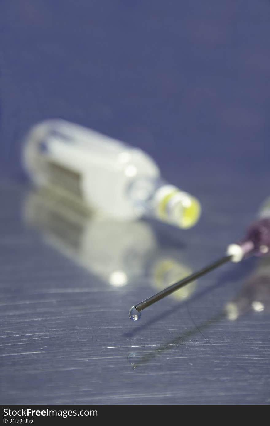 Syringe with sharp needle and droplet in shallow dof with ampule in background. Syringe with sharp needle and droplet in shallow dof with ampule in background