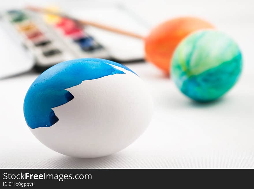 A half painted blue easter egg with color easter eggs, brush and watercolor palette in background. A half painted blue easter egg with color easter eggs, brush and watercolor palette in background.