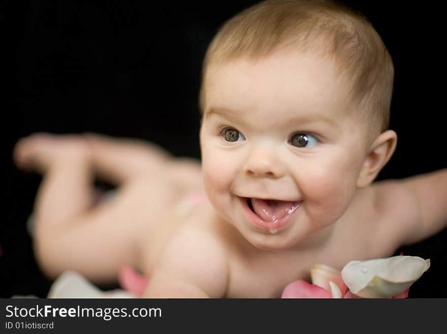 A naked baby smiling, on rose pedals,  against a black backdrop