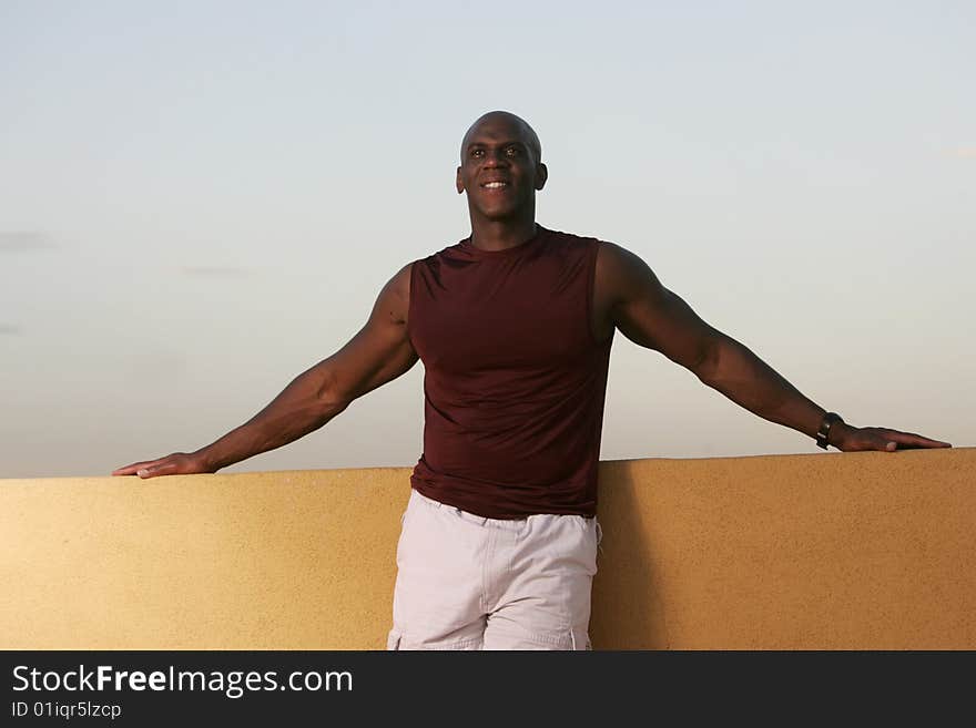 A male athlete with dark skin wearing a puple shirt relaxes out doors against a mustard wall and blue sky. A male athlete with dark skin wearing a puple shirt relaxes out doors against a mustard wall and blue sky