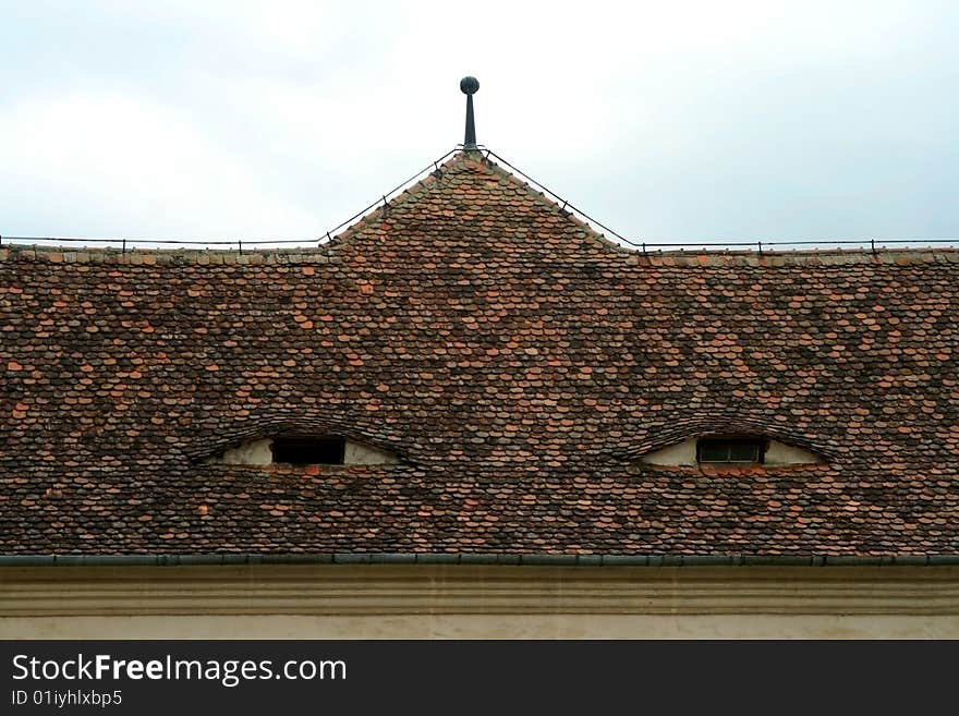 An old roof of tiles from a historic fortress watching sleepy through its windows, in Fagaras, Romania. An old roof of tiles from a historic fortress watching sleepy through its windows, in Fagaras, Romania.