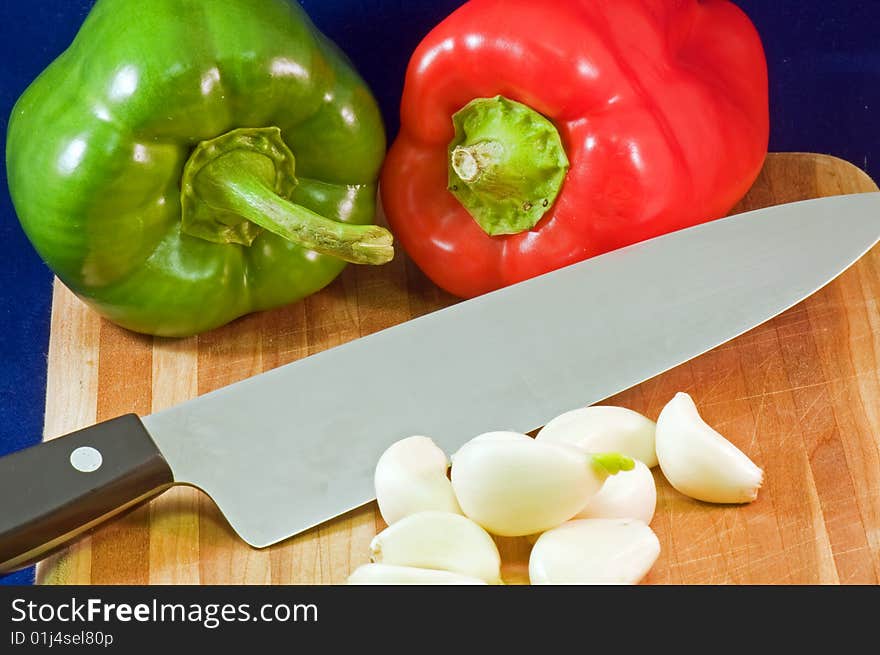 Whole red and green peppers on a wooden cutting board with garlic cloves and a large kitchen knife. Whole red and green peppers on a wooden cutting board with garlic cloves and a large kitchen knife.