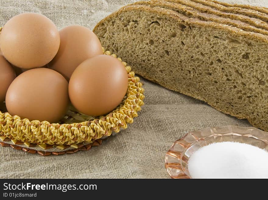 Many fresh rural eggs lying in a wattled small basket near to fresh bread and saltcellar on a cloth from a rough fabric. Many fresh rural eggs lying in a wattled small basket near to fresh bread and saltcellar on a cloth from a rough fabric