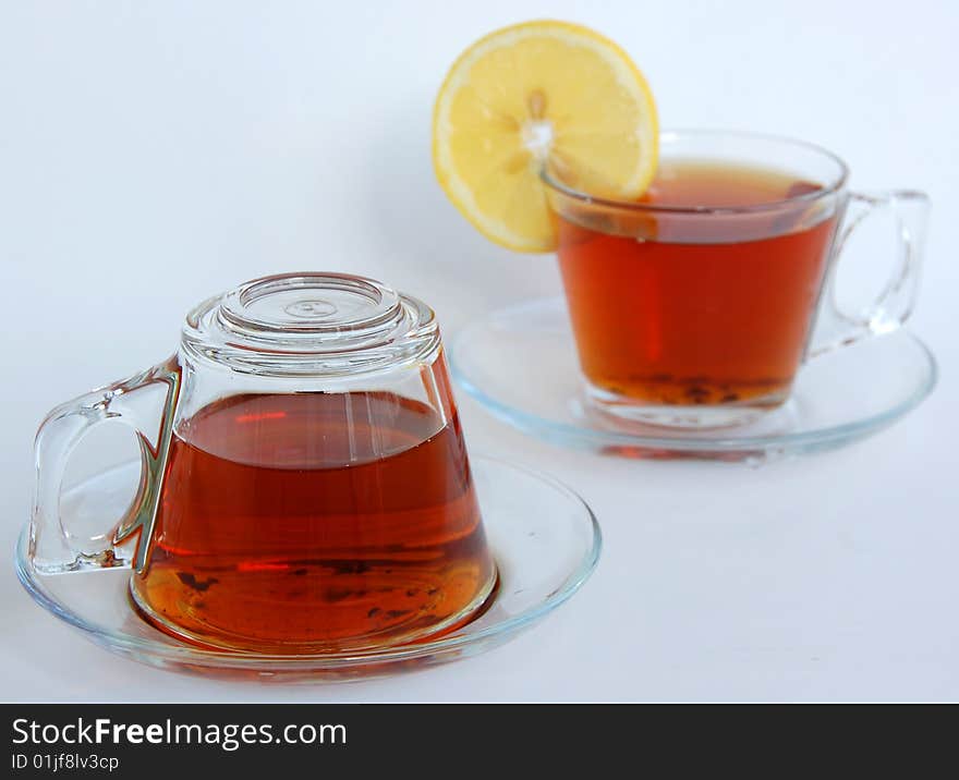 Two full glass teacups of modern design with handle and saucer, one upside-down, the other has a slice of yellow lemon. Two full glass teacups of modern design with handle and saucer, one upside-down, the other has a slice of yellow lemon.