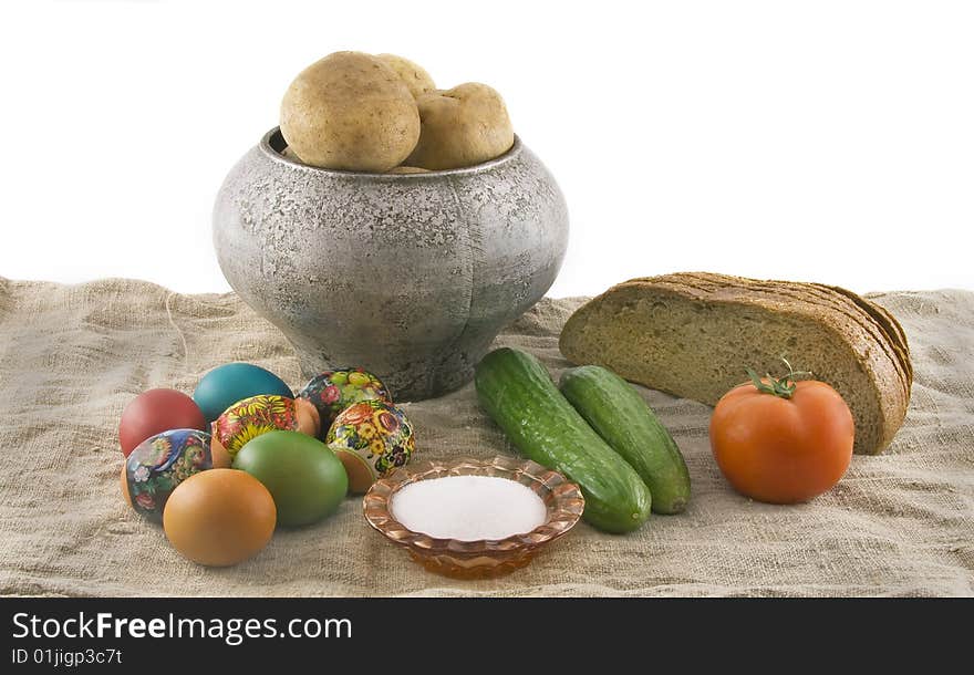 Still-life from vegetarian food. Fresh cucumbers, easter decorated rural eggs, a ripe tomato, fragrant rye bread, a boiled potato in a pig-iron kettle, a saltcellar, lying on sacking rough fabric. Isolated over white background