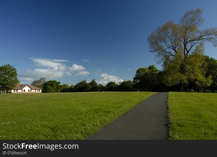 A path stretching away across an empty park with an almost clear blue sky in the background. Space for text in the sky or on the grass.