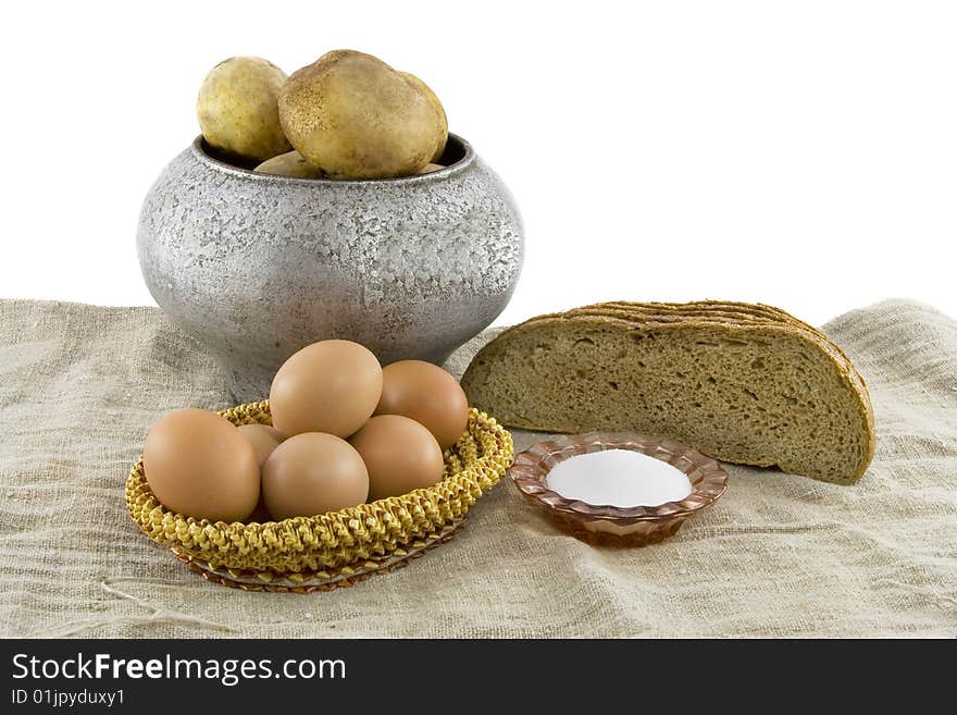 Still-life from vegetarian food. Fresh eggs lying in a wattled small basket, fragrant rye bread, a boiled potato in a pig-iron kettle, a saltcellar on a rough fabric