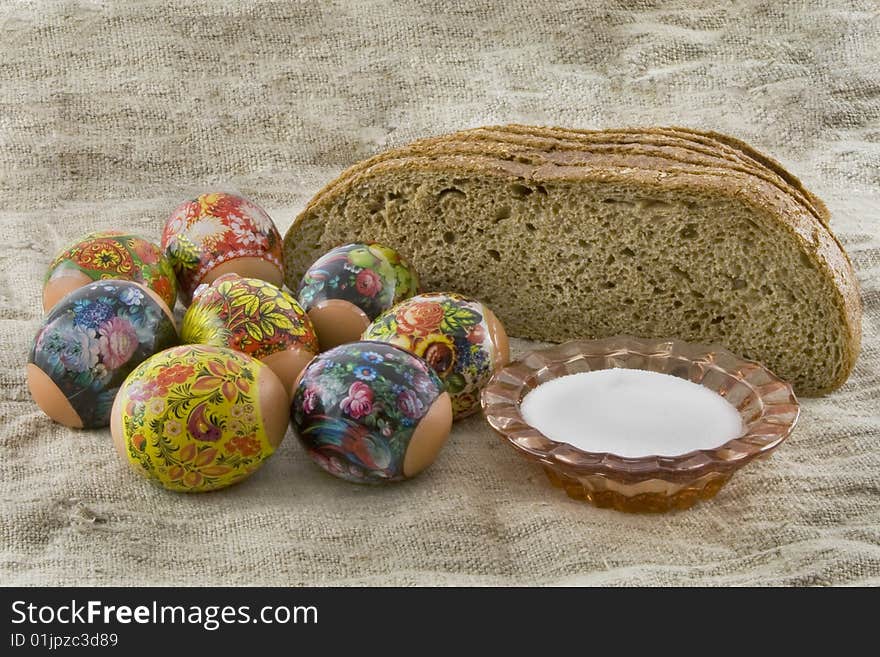 Many fresh easter rural decorated eggs lying in a wattled small basket near to fresh bread and saltcellar on a cloth from a rough fabric. Many fresh easter rural decorated eggs lying in a wattled small basket near to fresh bread and saltcellar on a cloth from a rough fabric