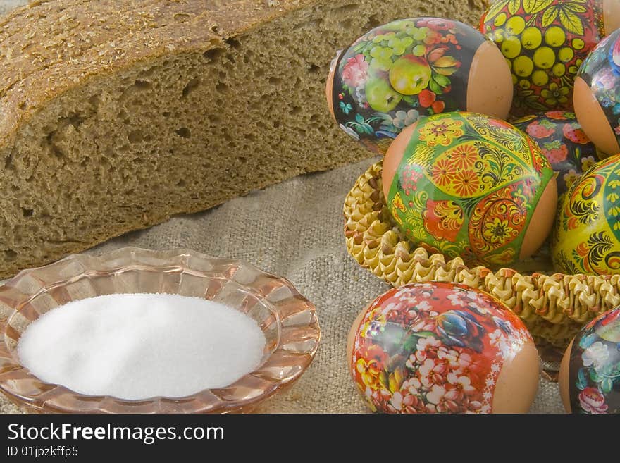 Many fresh easter rural decorated eggs lying in a wattled small basket near to fresh bread and saltcellar on a cloth from a rough fabric