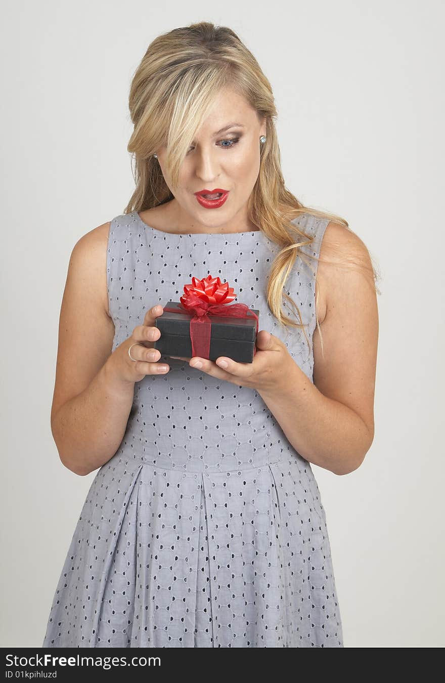 Portrait of a beautiful blonde woman with light blue eyes and classic make-up holding a gift box with excitement on grey background. Portrait of a beautiful blonde woman with light blue eyes and classic make-up holding a gift box with excitement on grey background