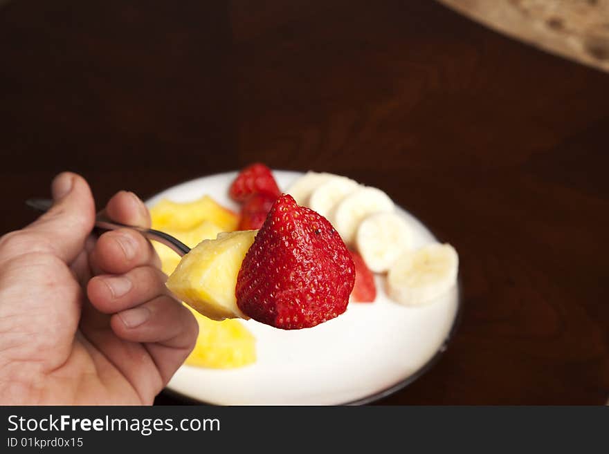 A strawberry and pineapple slice on a fork with a plate of fruit in the background. A strawberry and pineapple slice on a fork with a plate of fruit in the background.