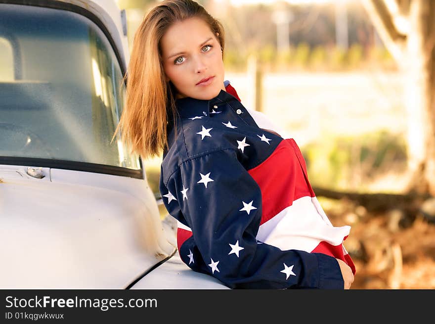 Attractive country girl in American flag shirt leans back against an old truck in rural america. Attractive country girl in American flag shirt leans back against an old truck in rural america