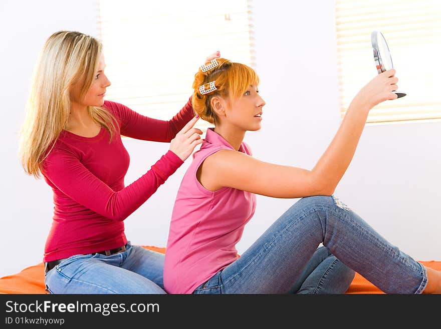 Young woman styling hair her friend. Red-haired woman with curlers on head holding hand glass. Side view. Young woman styling hair her friend. Red-haired woman with curlers on head holding hand glass. Side view.