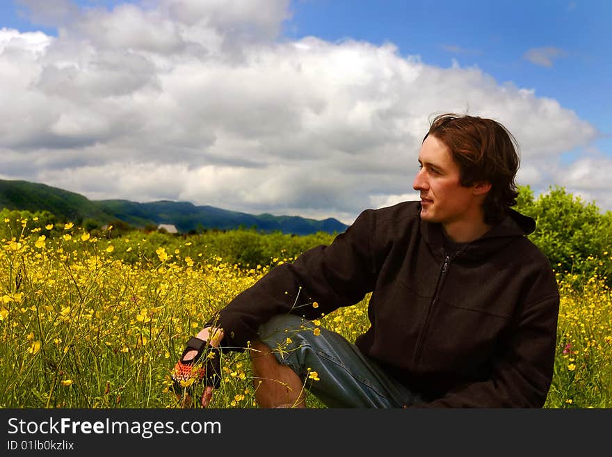 Young caucasian man wearing black sweater and sport gloves sits on spring meadow full of yellow flowers in sunny weather. Young caucasian man wearing black sweater and sport gloves sits on spring meadow full of yellow flowers in sunny weather