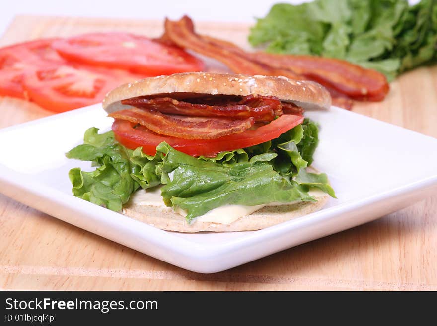 Bacon, lettuce and tomato sandwich with a flat bun on a white plate with ingredients in the background. Bacon, lettuce and tomato sandwich with a flat bun on a white plate with ingredients in the background