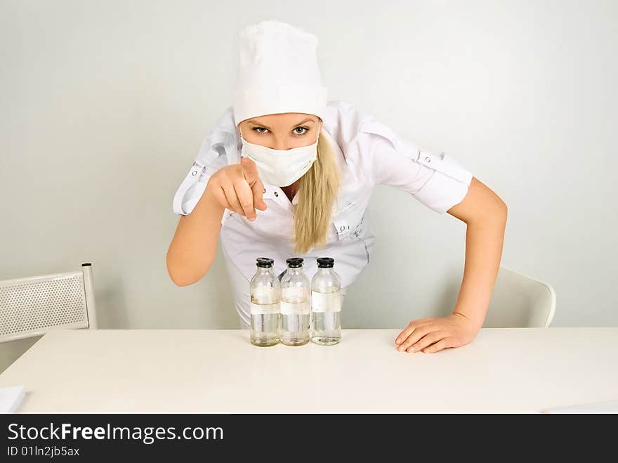 Portrait of a young doctor with medications sitting by the table in her office and pointing at us with a finger. Portrait of a young doctor with medications sitting by the table in her office and pointing at us with a finger