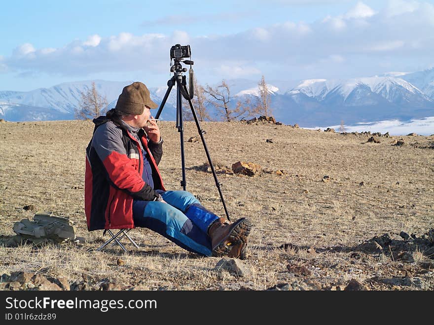 Old man sitting and in the mountains with camera standing on tripod. Old man sitting and in the mountains with camera standing on tripod