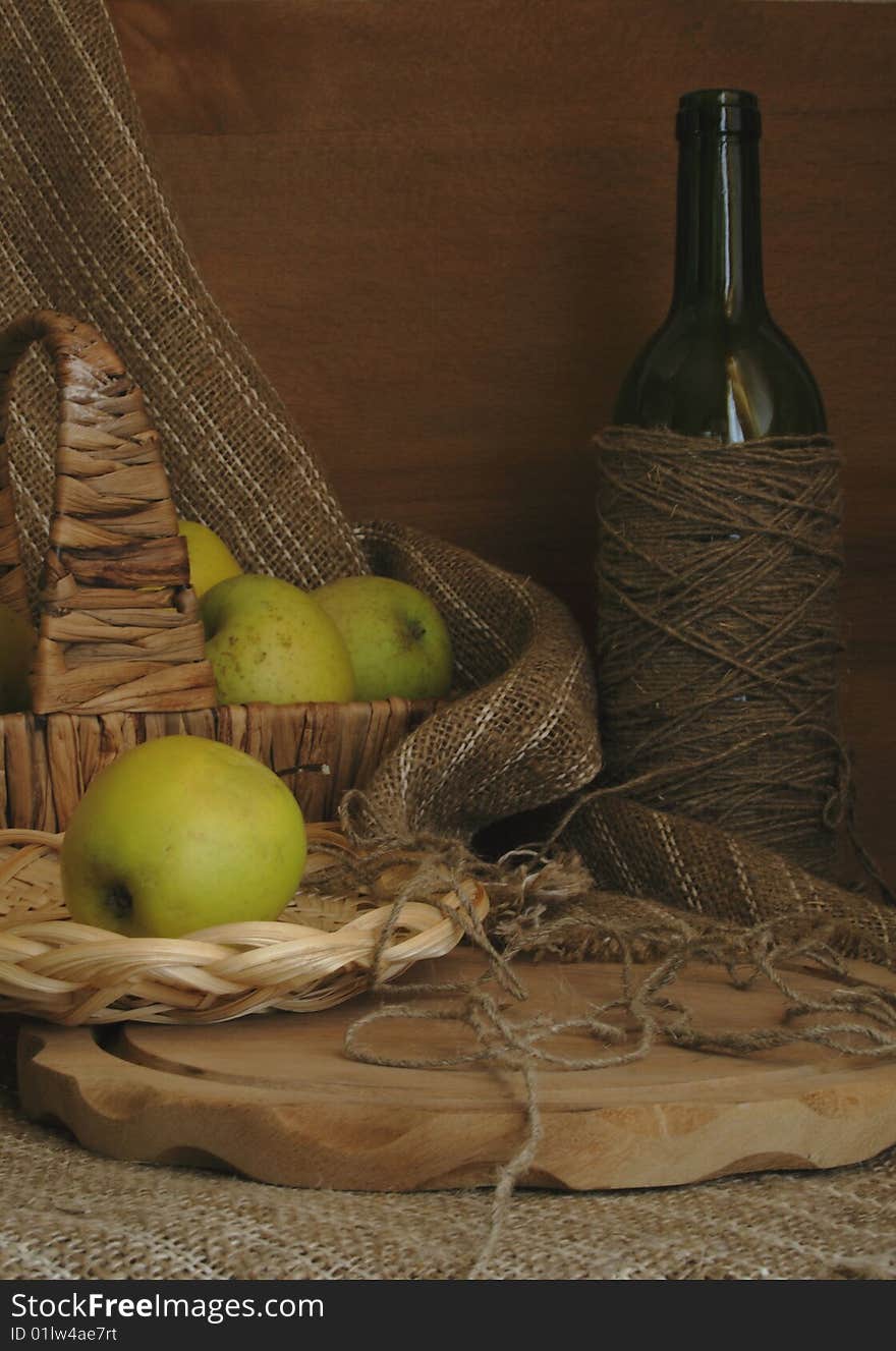 Green apples in a basket, a bottle, a chopping board against a homespun drapery. Green apples in a basket, a bottle, a chopping board against a homespun drapery