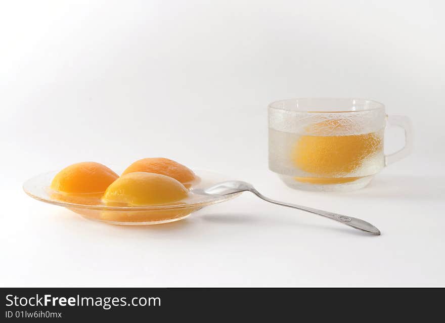 Segments of tinned peaches on a glass saucer with a dessertspoon and a cup with a peach and a syrup. Segments of tinned peaches on a glass saucer with a dessertspoon and a cup with a peach and a syrup