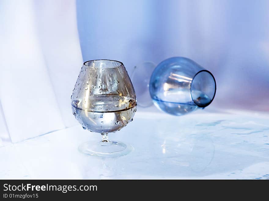 Two wineglasses, one stands the second lies,  on wet glass. Both with a liquid.