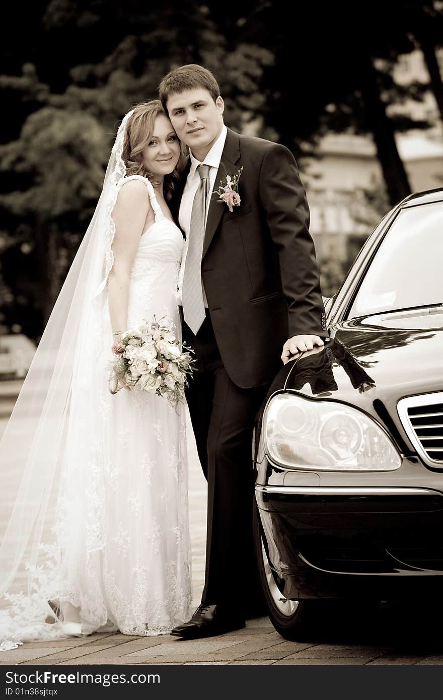 On the image there is bride and bridegroom. They are by car. On the image there is bride and bridegroom. They are by car.