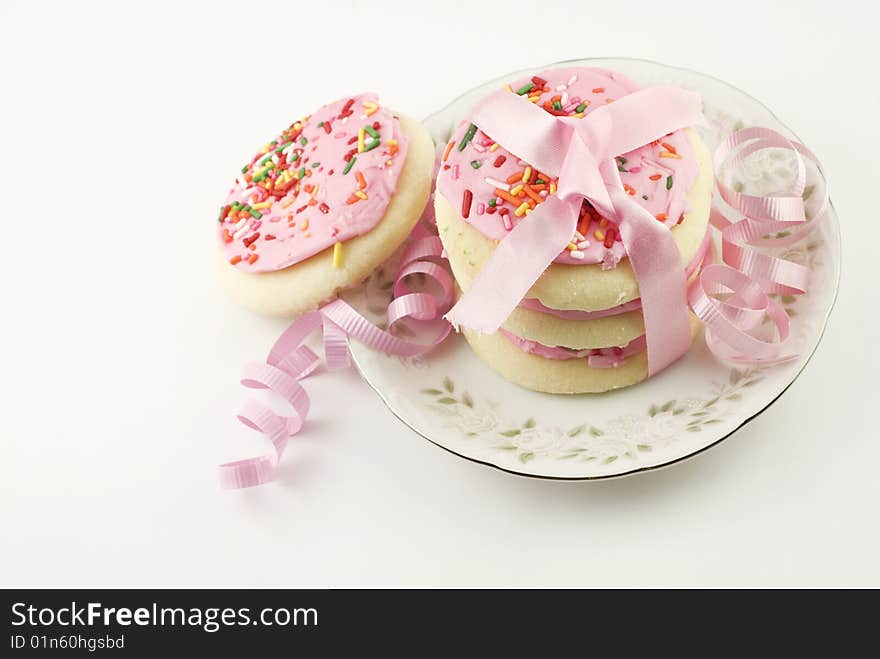 A plate of frosted pink sugar cookies with sprinkles in a stack tied with pink ribbon, isolated on white background with copy space. A plate of frosted pink sugar cookies with sprinkles in a stack tied with pink ribbon, isolated on white background with copy space