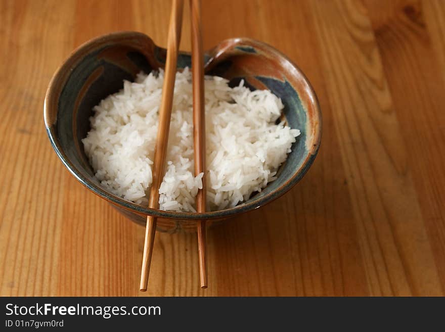 A bowl of steamed rice with wooden chopsticks through holes of the bowl on a wooden surface. A bowl of steamed rice with wooden chopsticks through holes of the bowl on a wooden surface.