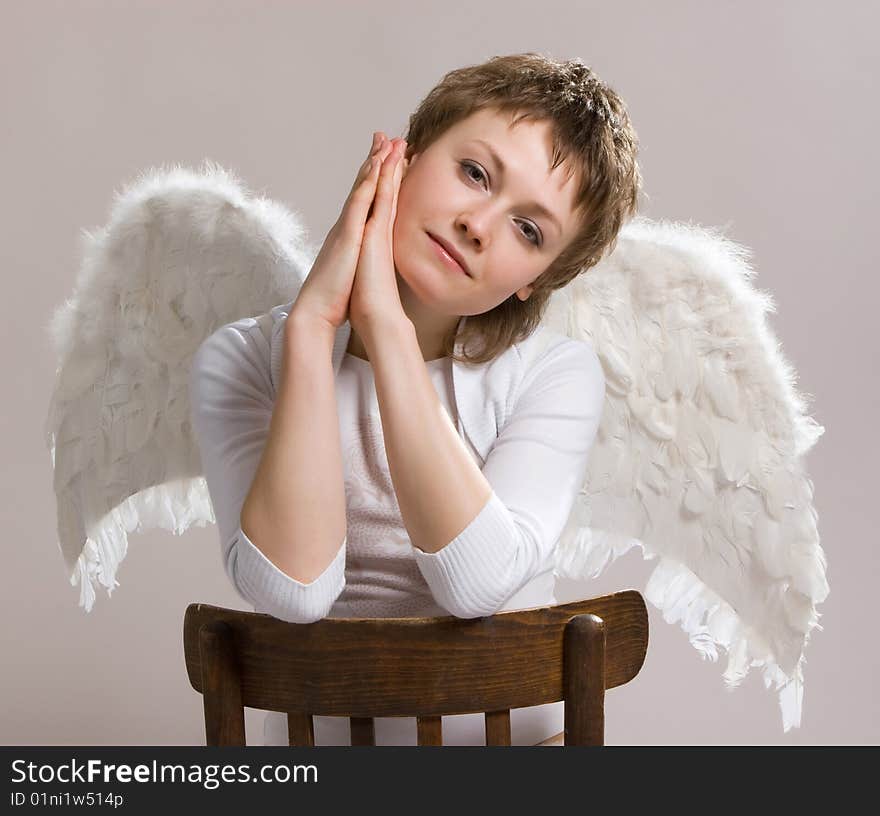 Silhouette of a pensive girl in white clothes on the light background sitting on the chair with wings of an angel behind her back. Silhouette of a pensive girl in white clothes on the light background sitting on the chair with wings of an angel behind her back