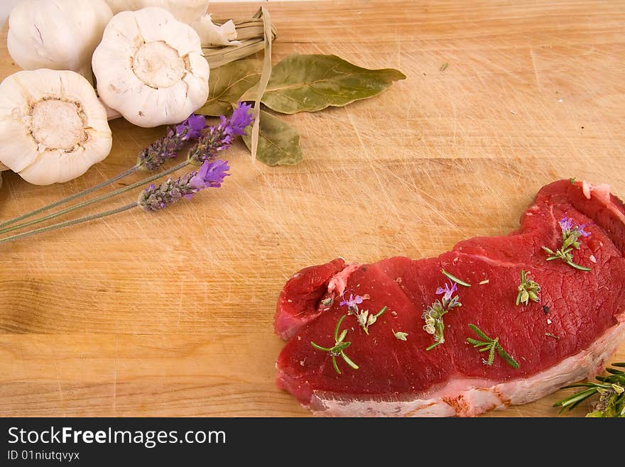 Two beef steaks, and a set of Mediterranean ingredients over a wooden board ready to be chopped and used to flavour a meal. Two beef steaks, and a set of Mediterranean ingredients over a wooden board ready to be chopped and used to flavour a meal