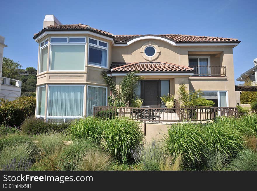 House with lots of shrubs in front. Blue sky. Two levels with yard of trees and shrubs. Circular window in front. Railings. Large picture windows.