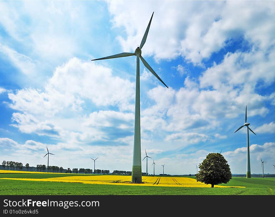 Wind turbines among rapeseed field and green meadows against a cloudy blue sky. Wind turbines among rapeseed field and green meadows against a cloudy blue sky