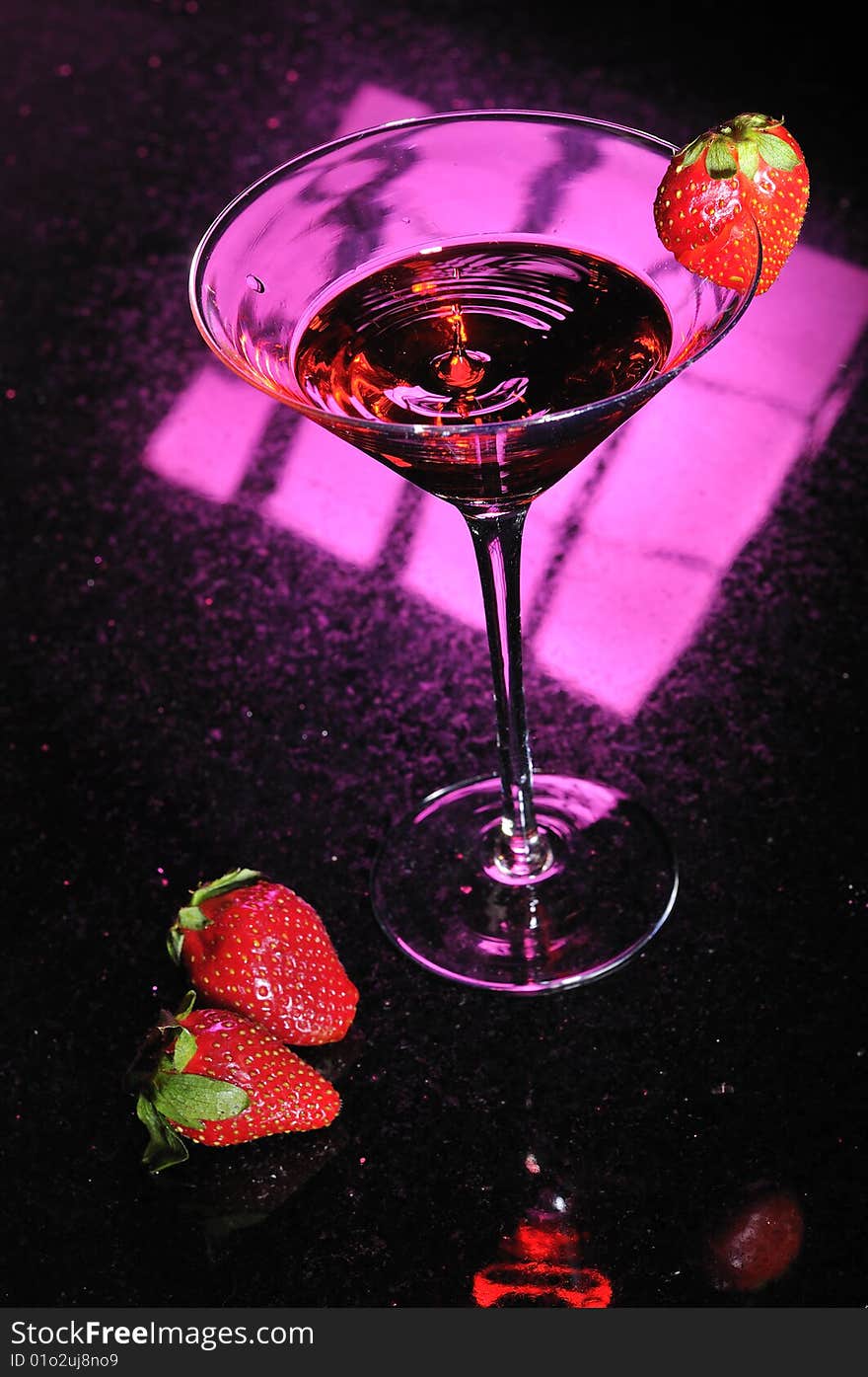 A martini glass with red liquid splashing up in center and a strawberry placed on side with two strawberries next to glass. A martini glass with red liquid splashing up in center and a strawberry placed on side with two strawberries next to glass