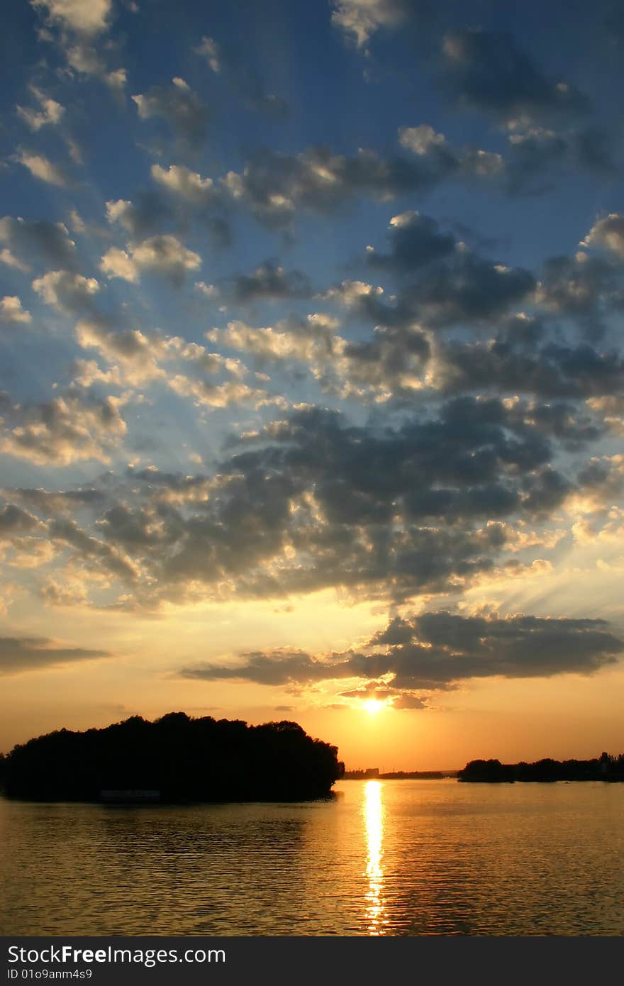 A bright sunset in a still evening deep in a high sky behind the river with a dark strip of trees