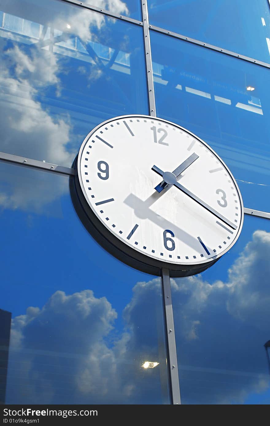 Picture of a clock on glass wall reflecting clouds and blue sky