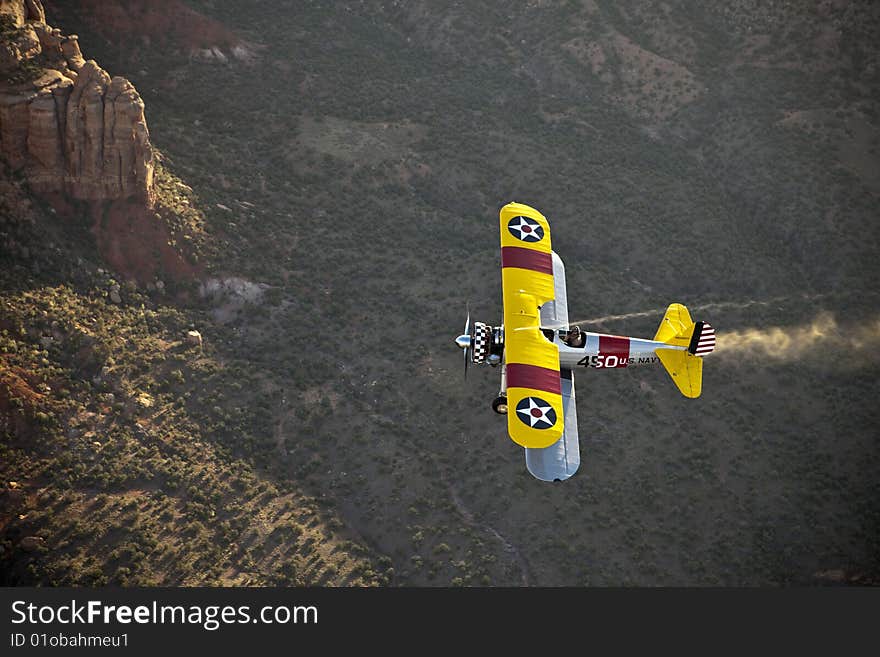 Yellow 1942 steerman flying over rugged desert with smoke from over head. Yellow 1942 steerman flying over rugged desert with smoke from over head