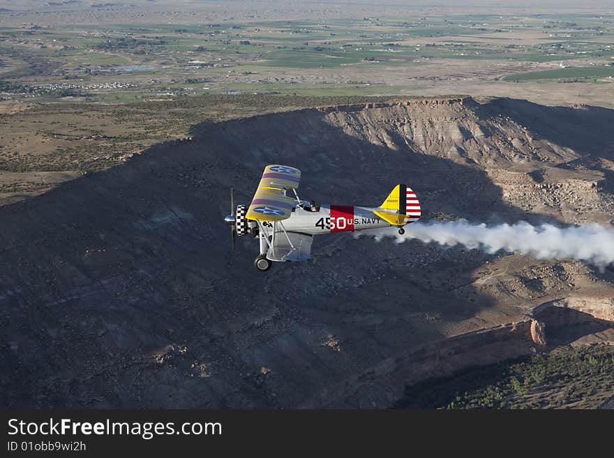 Yellow 1942 steerman flying over rugged desert with smoke from over head. Yellow 1942 steerman flying over rugged desert with smoke from over head