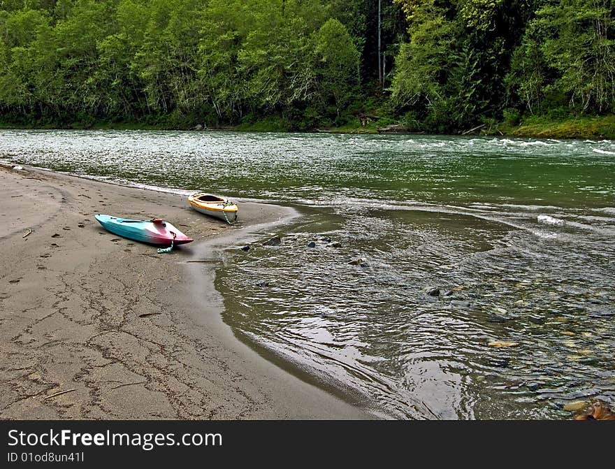 This peaceful photo is of two kayaks laying on a sandy river bed with the gorgeous river and mountains in the scene. This peaceful photo is of two kayaks laying on a sandy river bed with the gorgeous river and mountains in the scene.