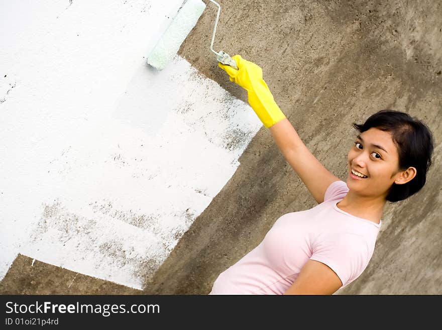 Independent asian young woman painting exterior wall of her house with a white colored paint, using a roller and gloves on hands. Independent asian young woman painting exterior wall of her house with a white colored paint, using a roller and gloves on hands